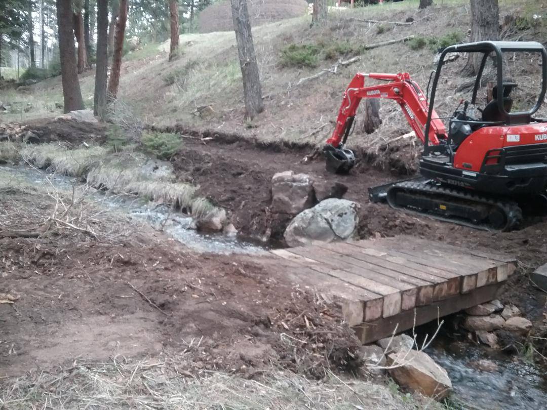 Compact excavator working a muddy drainage beside a small creek and timber bridge