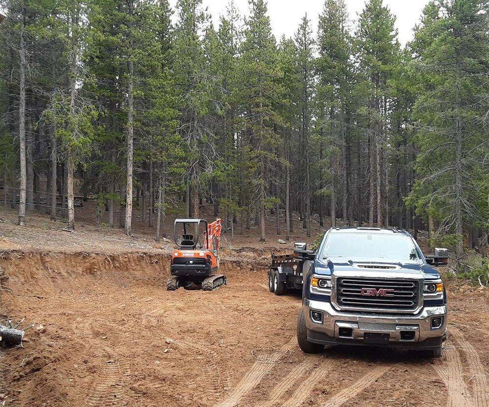 Orange excavator and a GMC truck on a freshly cut dirt driveway through pine forest