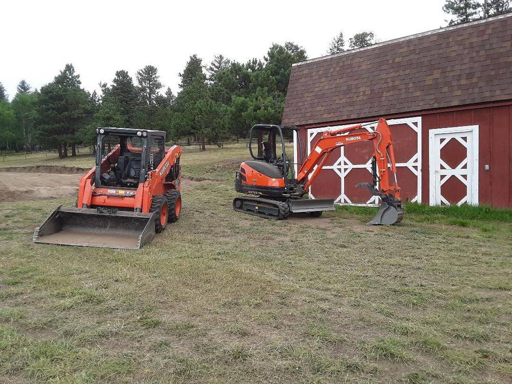 Kubota skid steer and compact excavator parked beside a red barn in the pines