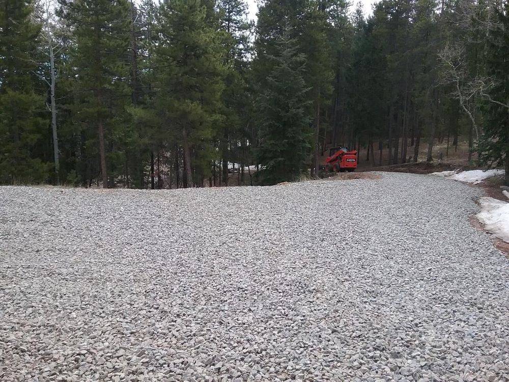 Wide gravel pad in a pine forest with an excavator in the background and snow at the edge