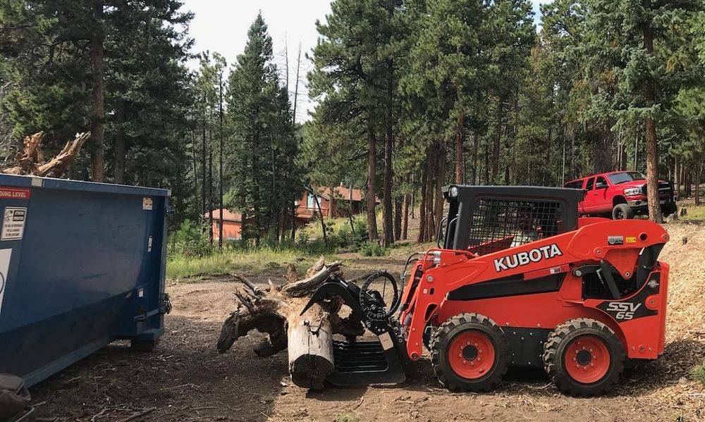 Kubota skid steer with a grapple moving a stump beside a dumpster